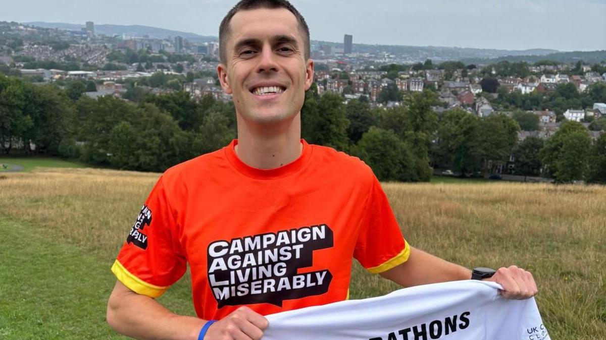 A smiley man in a bright orange sports shirt stands on a grass field with the city spread out behind him