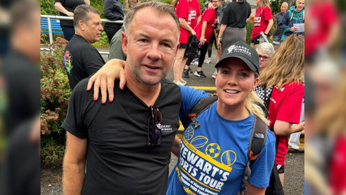 Marcus Stewart and his wife smiling with their arms around each other. She is wearing a black cap and a blue shirt that says 'Michael's Sports Tour'