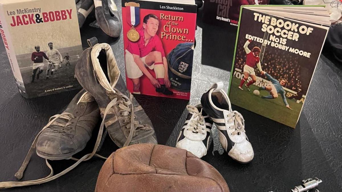 Two pairs of old football boots behind a deflated old football. Behind the boots are three books, one titled 'Jack & Bobby', the middle 'Return of the Clown Prince' and the one on the right 'The Book of Soccer No14, edited by Bobby Moore'.