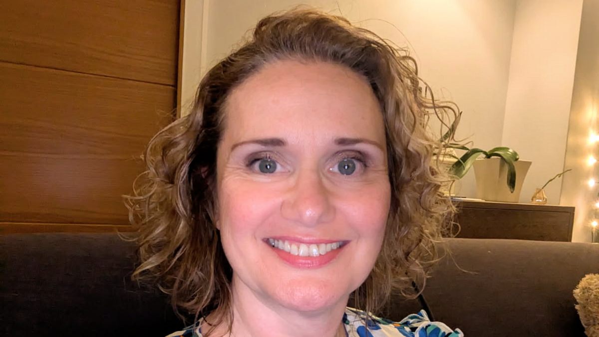 Emma Taylor is smiling at the camera, in front of a cupboard and a white wall and is sat on a sofa.