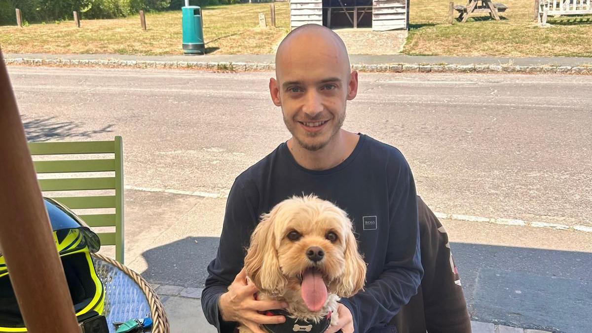 A young man with a bald head outdoors holding a shaggy small dog with its tongue out