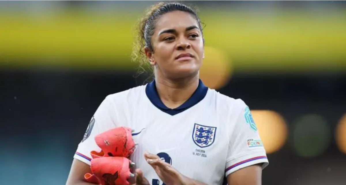 Jess Carter playing for England in a white and navy blue shirt. She looking into the distance while clapping with her pink football boots tucked under her left arm.