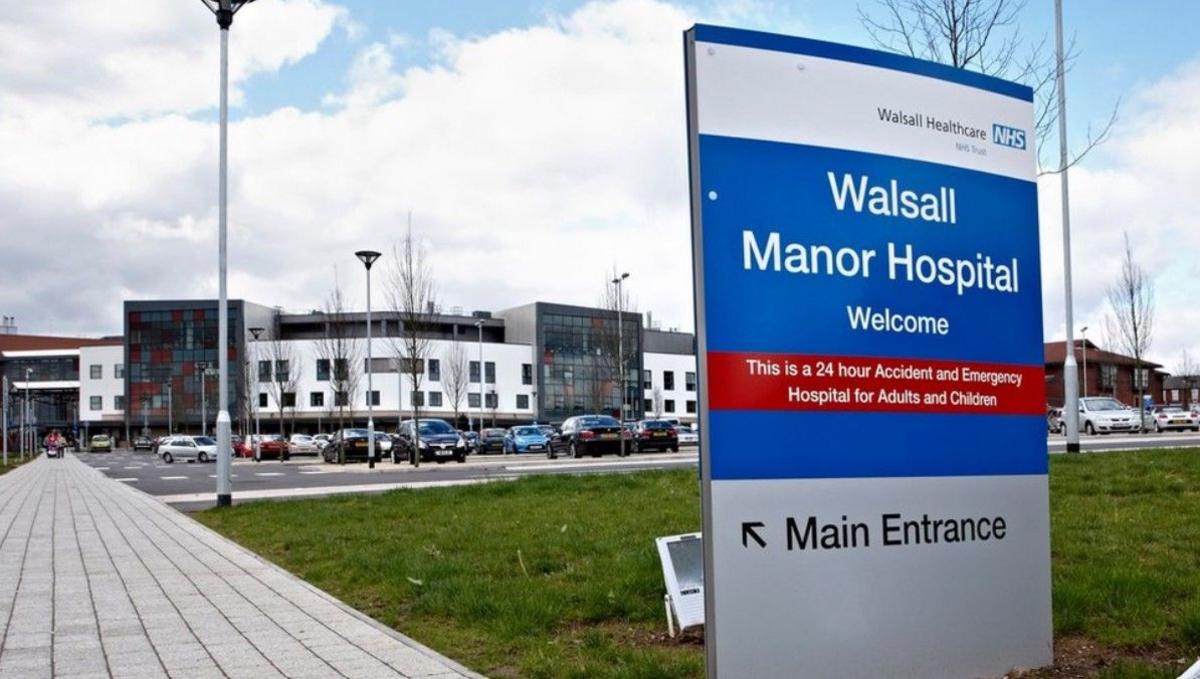 Walsall Manor Hospital in the distance - a white and grey building with a car park in front of it. There is a hospital sign in the foreground pointing people to the main entrance.