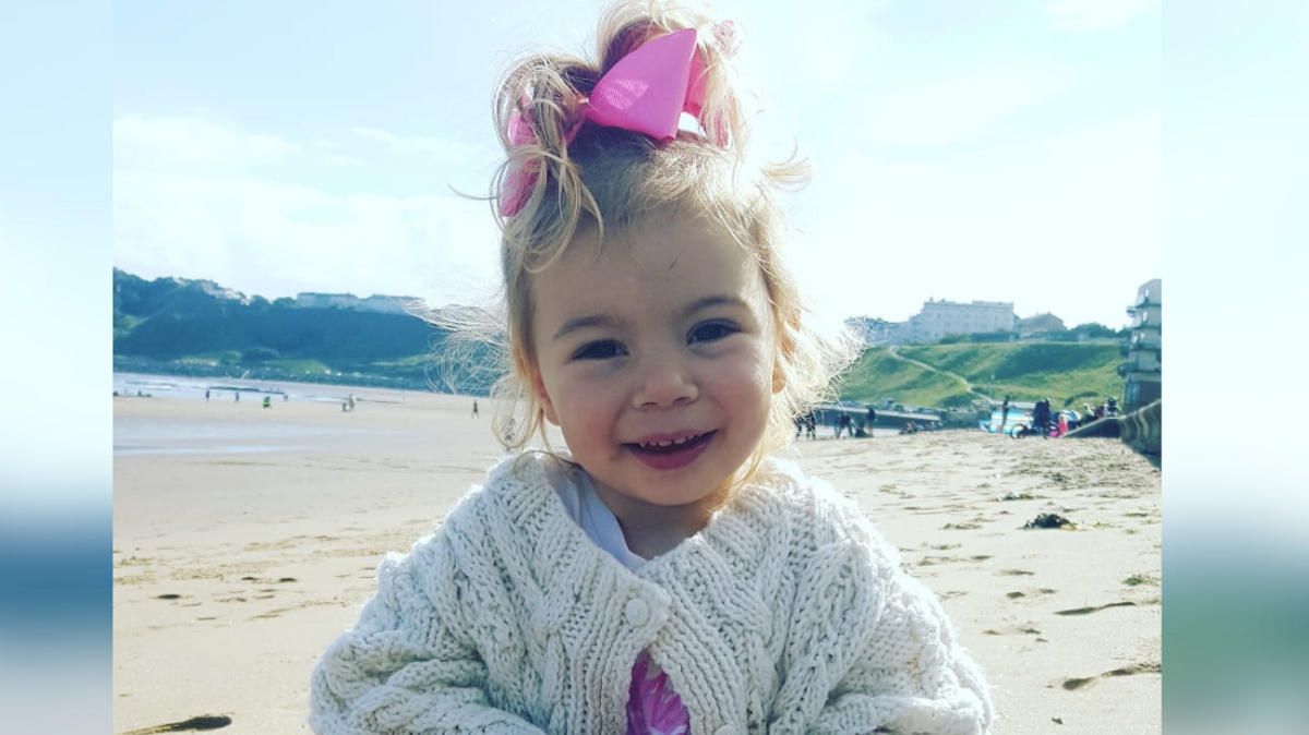 Thalia Johnson sits on the beach at Scarborough. She wears a white cardigan and pink leggings, with her blonde hair up in a pink bow.