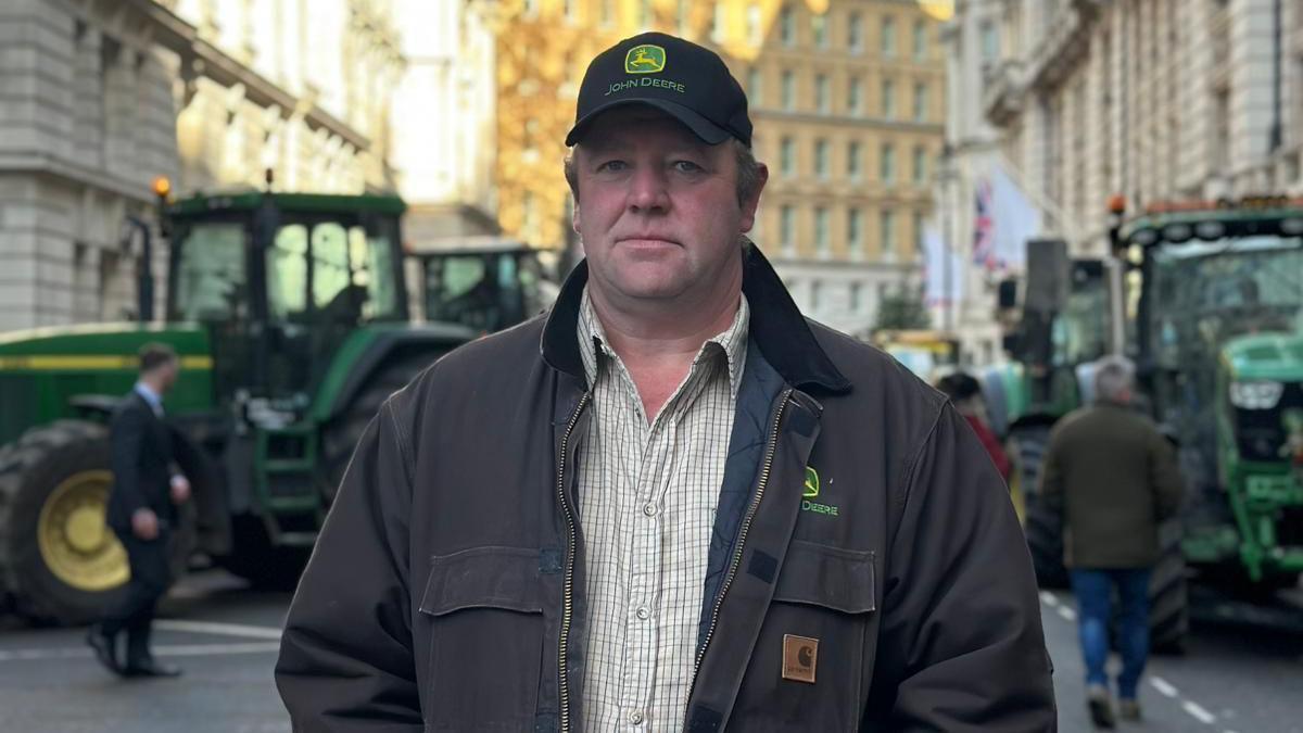 Dan stands in a London Street. He is wearing a black cap with a green logo, a white shirt with thin green lines and a dark coloured jacked. Tractors can be seen in the background. 