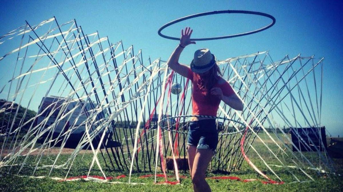 Lowri dancing with a hula hoop around her waist.  She is wearing a red short-sleeved top and denim shorts and in a field with an art installation covered in streamers behind her.