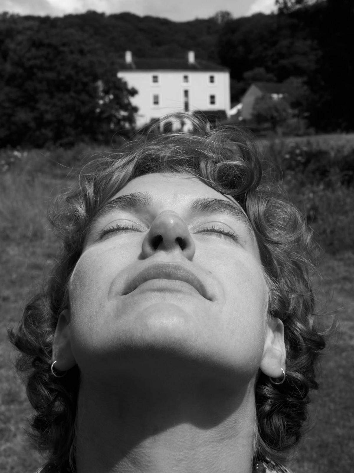 A black and white photo of a woman closing her eyes standing in front of a large country house 