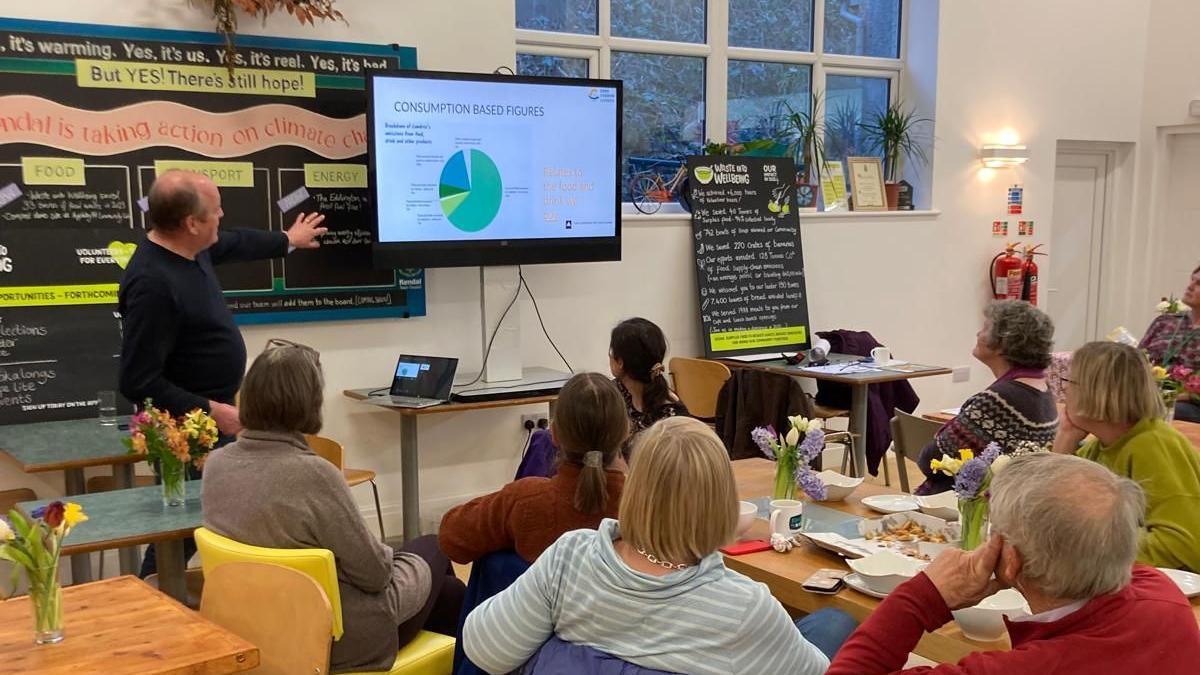Nick Lancaster pointing at a screen with a pie-chart. He is speaking to a group of people sat around a table. 