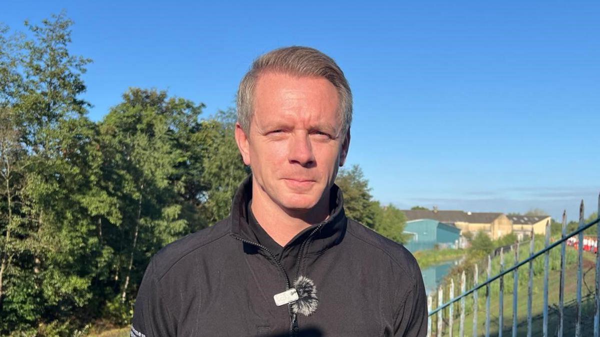 Marc Lidderth standing in sunshine by the canal. He has grey, black hair with a side parting and is slightly smiling at the camera and wearing a black jacket.