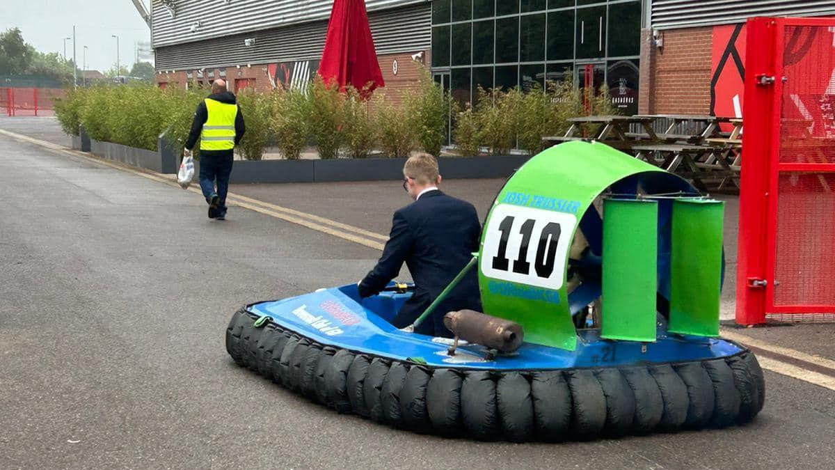 A photo taken from behind showing a young teenage boy driving a blue and green hovercraft with a red brick building in the background