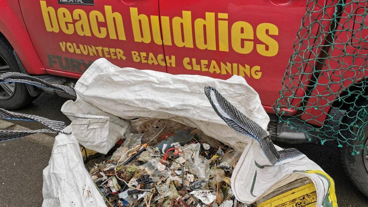 A big white bag of rubbish in front of a red van with yellow writing which reads beach buddies volunteer beach cleaning.