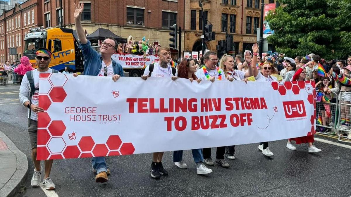 A group of people dressed in rainbow-themed clothing carry a banner reading "Telling HIV stigma to buzz off"