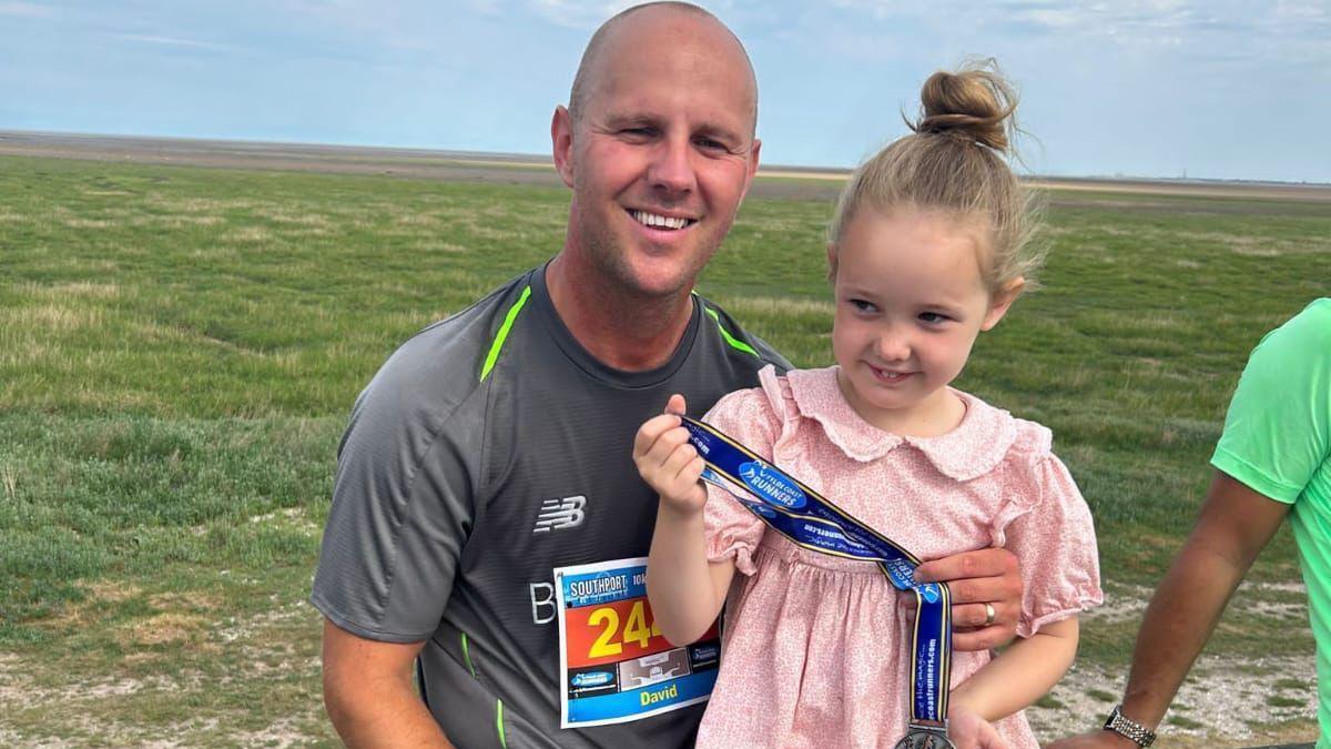 David and his daughter Elsie in 2023 after David had completed the Southport 10k. Elsie is wearing a pink dress with her hair in a bun and holding David's medal while David wears a grey running top and smiles at the camera.