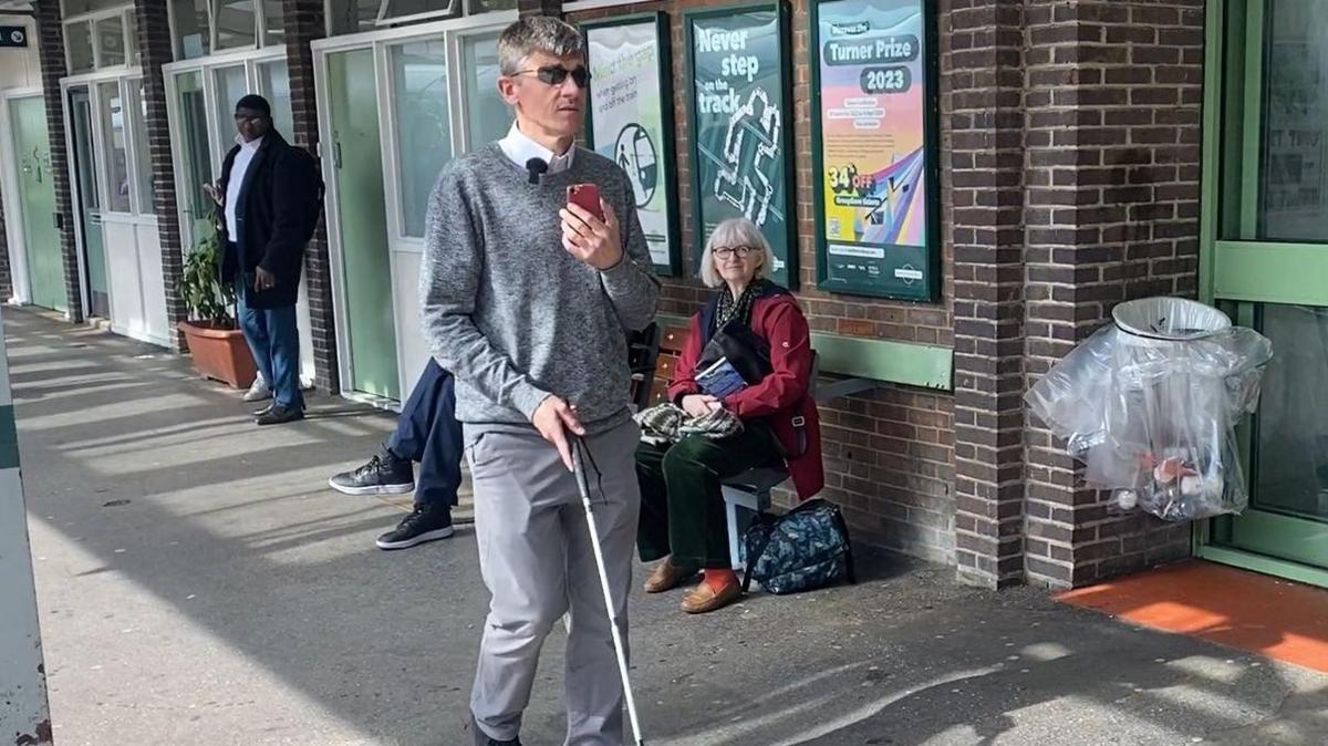 Dave Smith walking on a rail station platform with his white stick