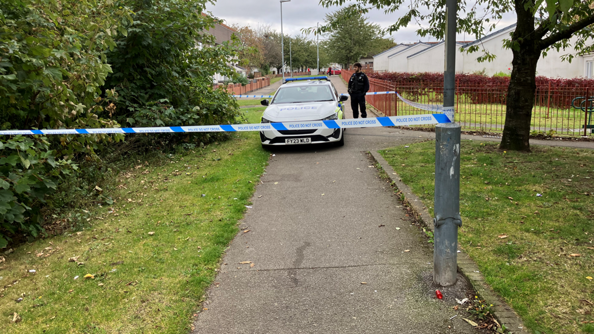 A photo of Hills Drive cordoned off with police tape. A police officer is standing behind the tape.