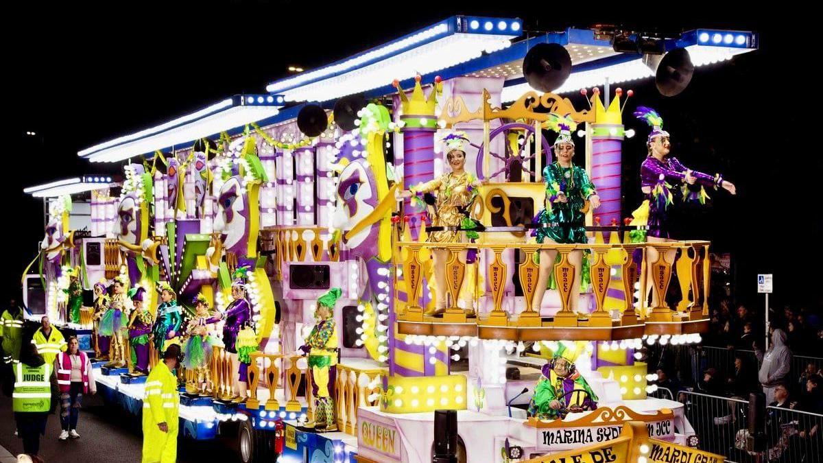 A brightly illuminated carnival float, showing purple and yellow colours and costumed performers around it.