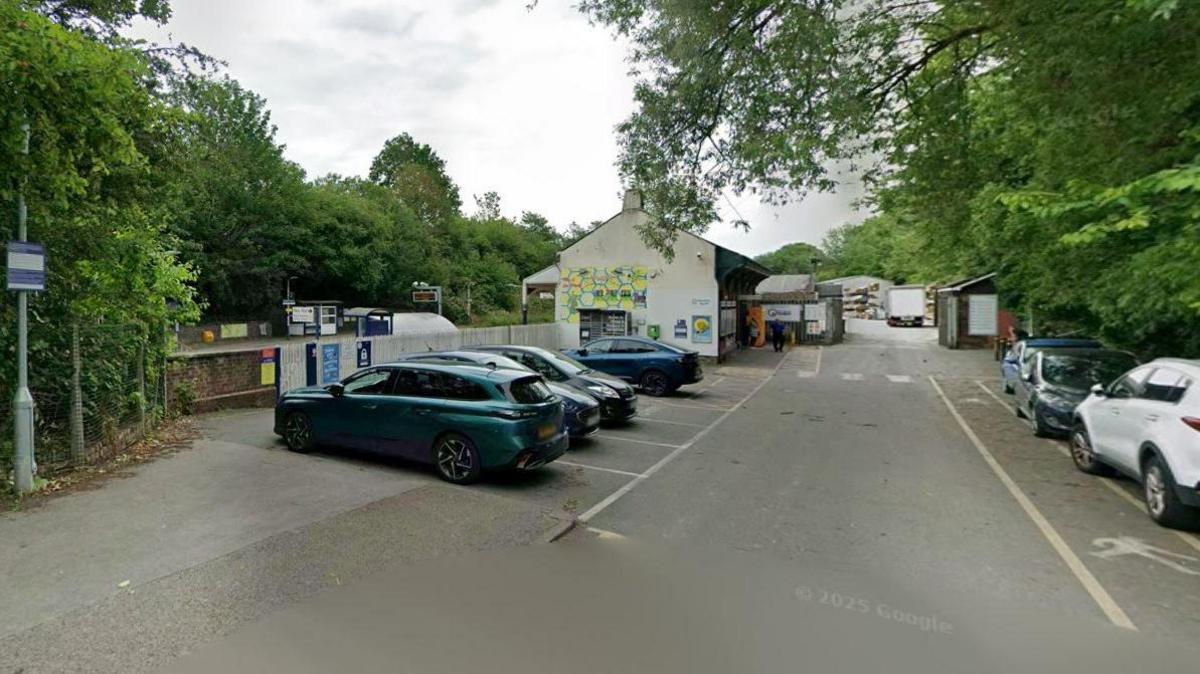 There are a number of cars parked up outside Reddish North railway station. The railway tracks are out of view, partially covered by a white fence. 