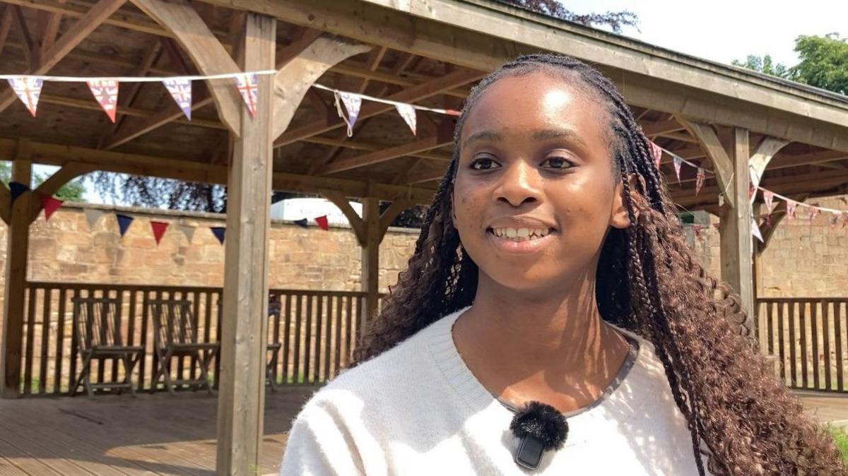 A girl with long braided black hair, wearing a white jumper, is standing in front of a wooden pavilion with Union Jack bunting around the outside. 