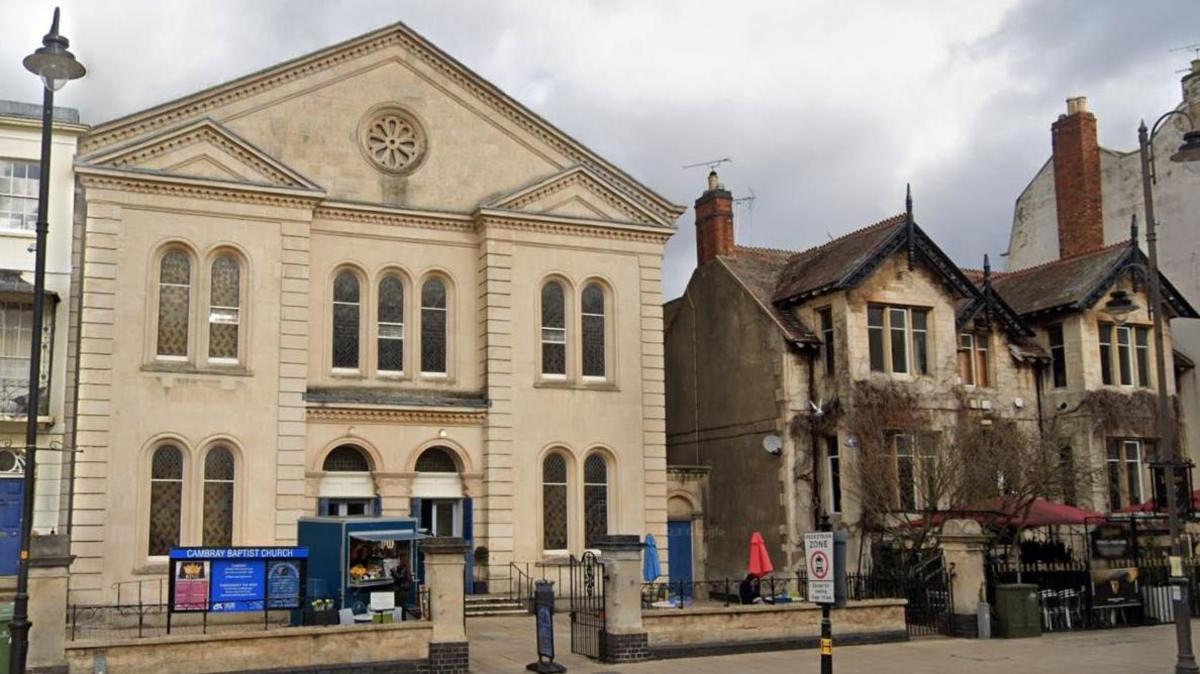 Cambray Baptist Church, an ornate, light coloured stone building with a pointy roof next to a cafe built with stone bricks on a town centre street