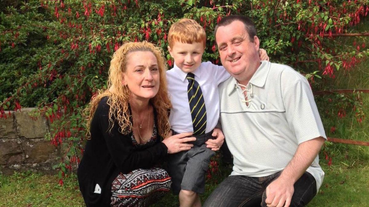 Smiling parents and their young boy smile for the camera on the boy's first day of school - the mum has strawberry blonde hair and is wearing a black top and multi-coloured dress. The dad is wearing a light top, jeans and has close cropped dark hair. The boy has reddish hair, a green tie and grey shorts.