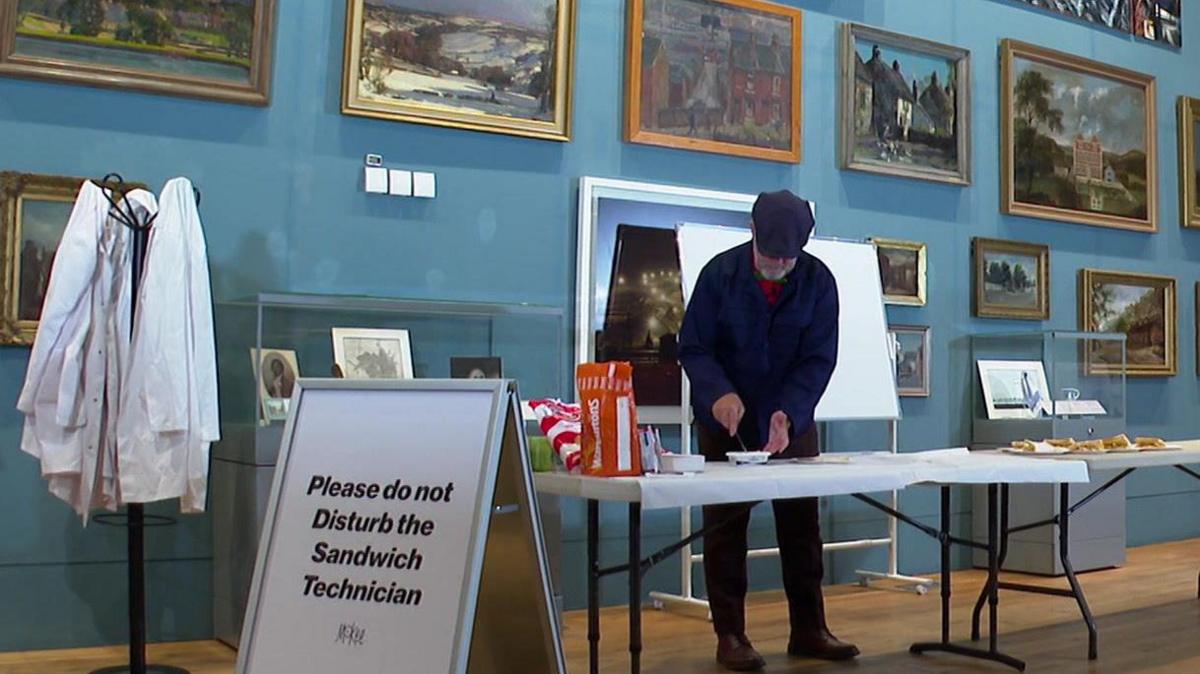 A man in a blue flat cap makes crisp sandwiches at white table in an art gallery. In front of him is an A-board that reads 'Please do not disturb the sandwich technician'.