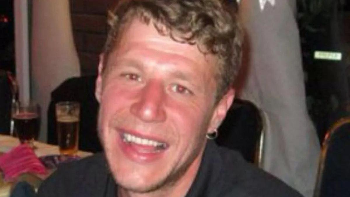 A young man smiles broadly at the camera. He has curly brown hair, a small earring and is wearing a black top. 