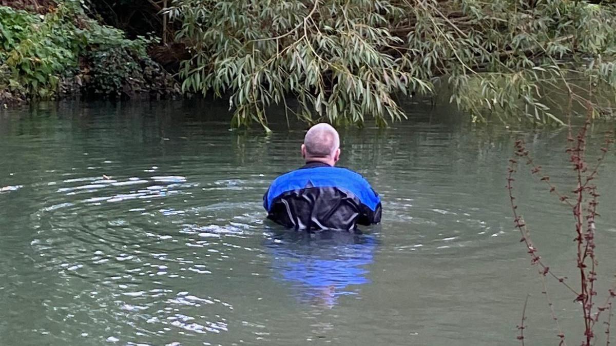 Back of a man in the river with water halfway up his chest, water is murky and a tree branch hangs low in front of him.