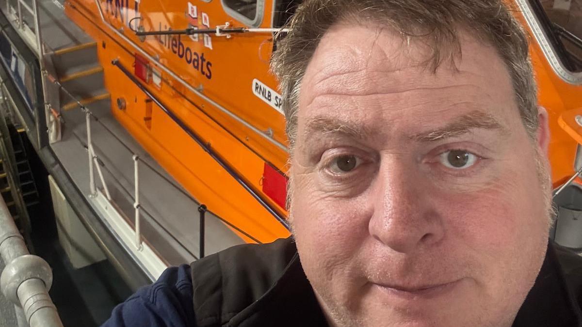 Michael England stands in front of an orange RNLI lifeboat inside Padstow RNLI's base.