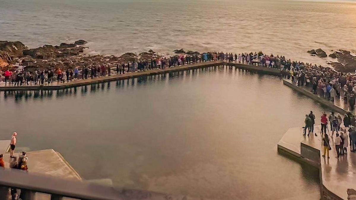 Hundreds of people gathered at La Vallette bathing pools on the seafront in Guernsey. It is a sunny day and the sea is relatively calm.