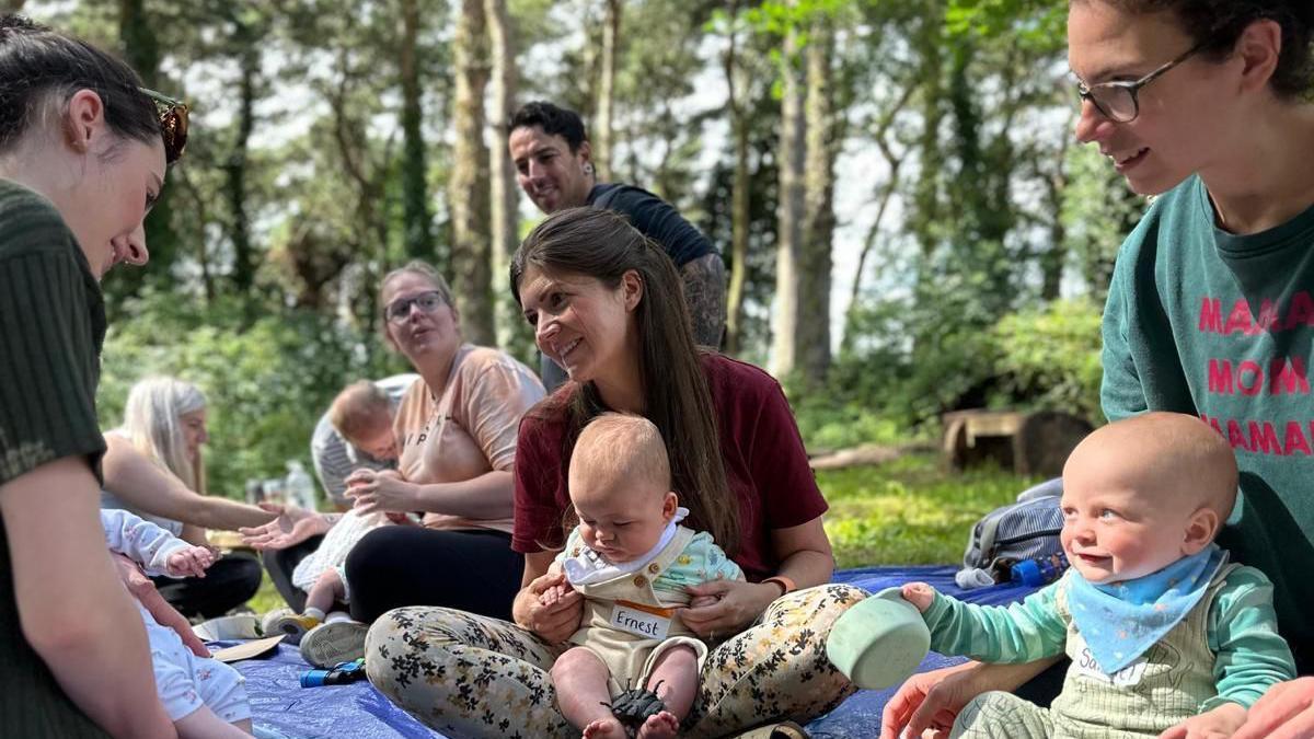 A group of parents and their babies sit in a park on a blue picnic blanket.