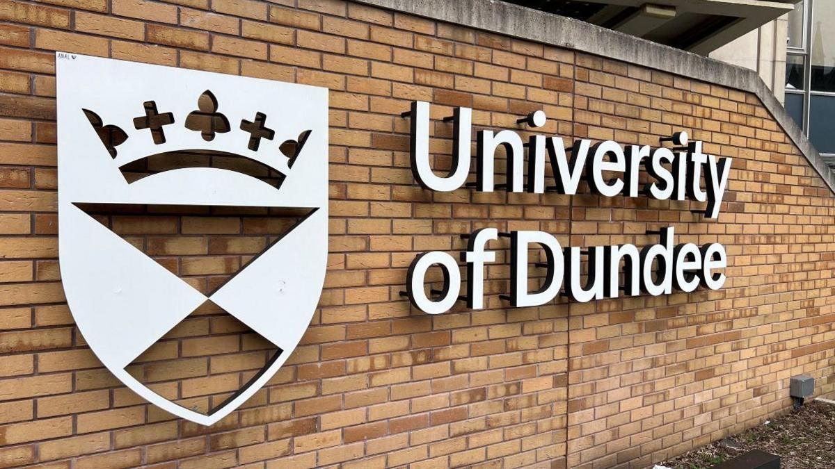 A white sign on a brick wall with a crest and the words University of Dundee