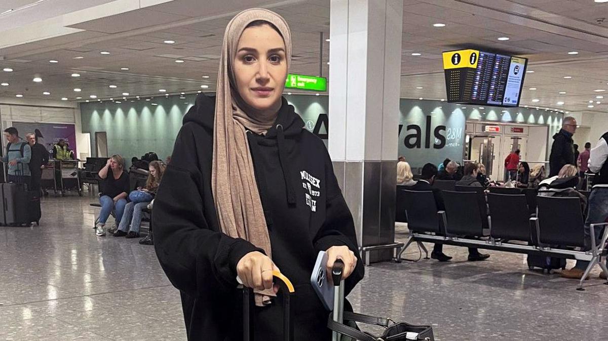 Alaa Ghalayini standing in Heathrow airport and looking straight at the camera. She is wearing a black hooded top, and a light gold coloured head scarf, gripping onto her luggage. Behind her people can be see sitting on airport seats, with a large arrival sign obscured behind a column. 