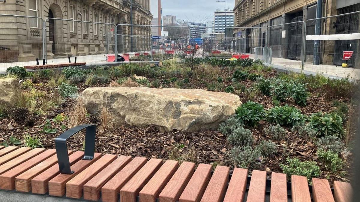 A new bench which has been installed in Bradford next to a new flower bed, shown with shrubs and rocks, with a street view in the background. Metal security fences and roadworks are in the background.