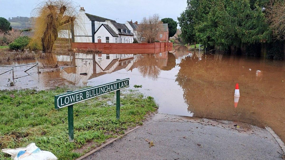 A road sign reading Lower Bullingham Lane, with pavement and grass area flooded with brown water. A house in the background is surrounded by a red brick wall, but the flooding extends past the house. 