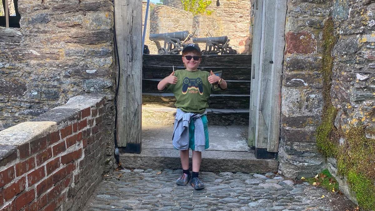 A young boy stands in an archway smiling at the camera with his thumbs up. He is wearing a cap, sunglasses, a green T-shirt and shorts with a blue top tied around his waist.