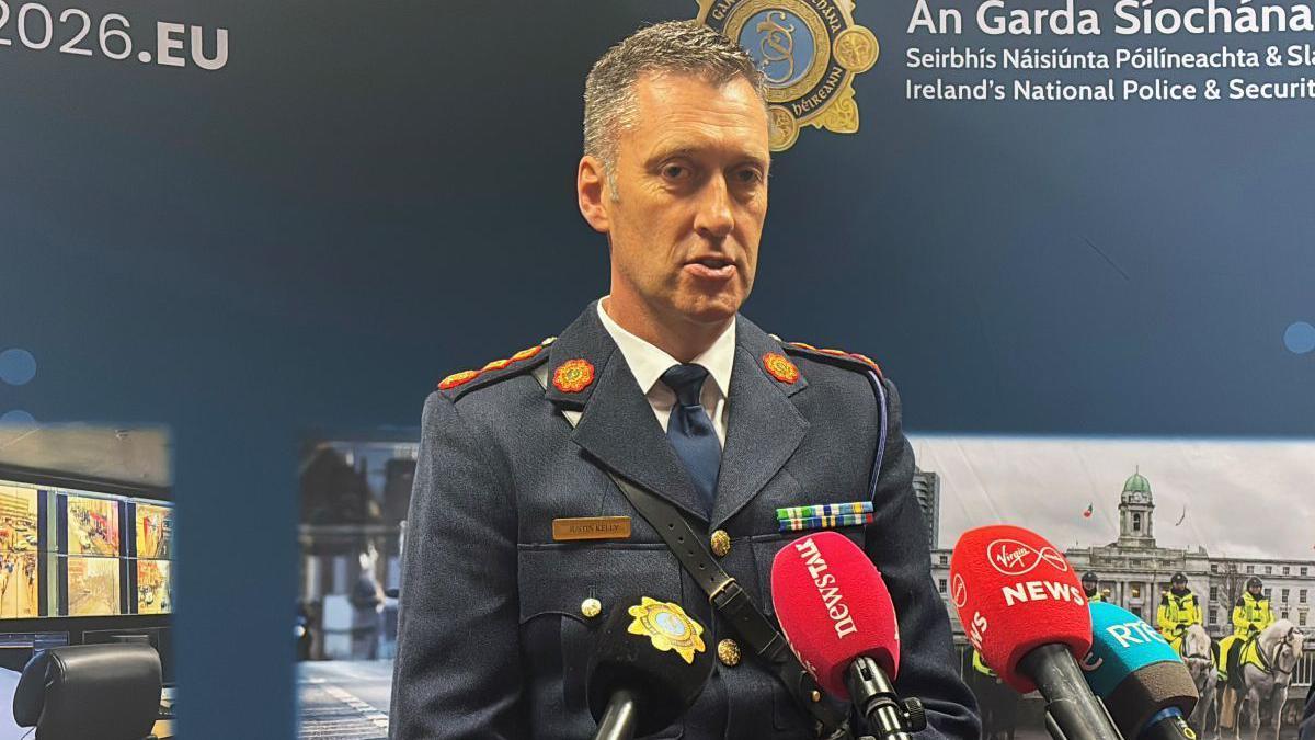 Garda officer wearing a blue uniform. He has short dark and grey hair and has mics in front of him. An Garda Siochana is on the background behind with police pictures.
