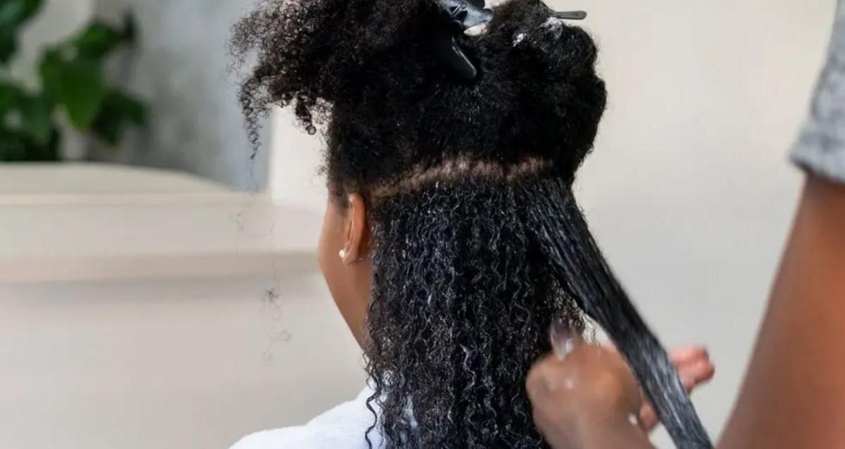 A woman with afro hair is sat with her back to the camera while a person who is out of vision is caring for her hair