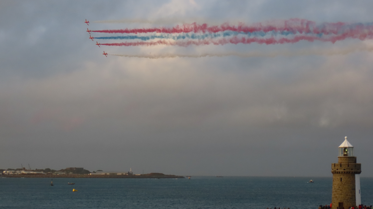 St Peter Port in Guernsey. The red arrows, a formation of red jets, are flying across the sky above the sea. There is a stone lighthouse to the right and a peninsula of land on the left.