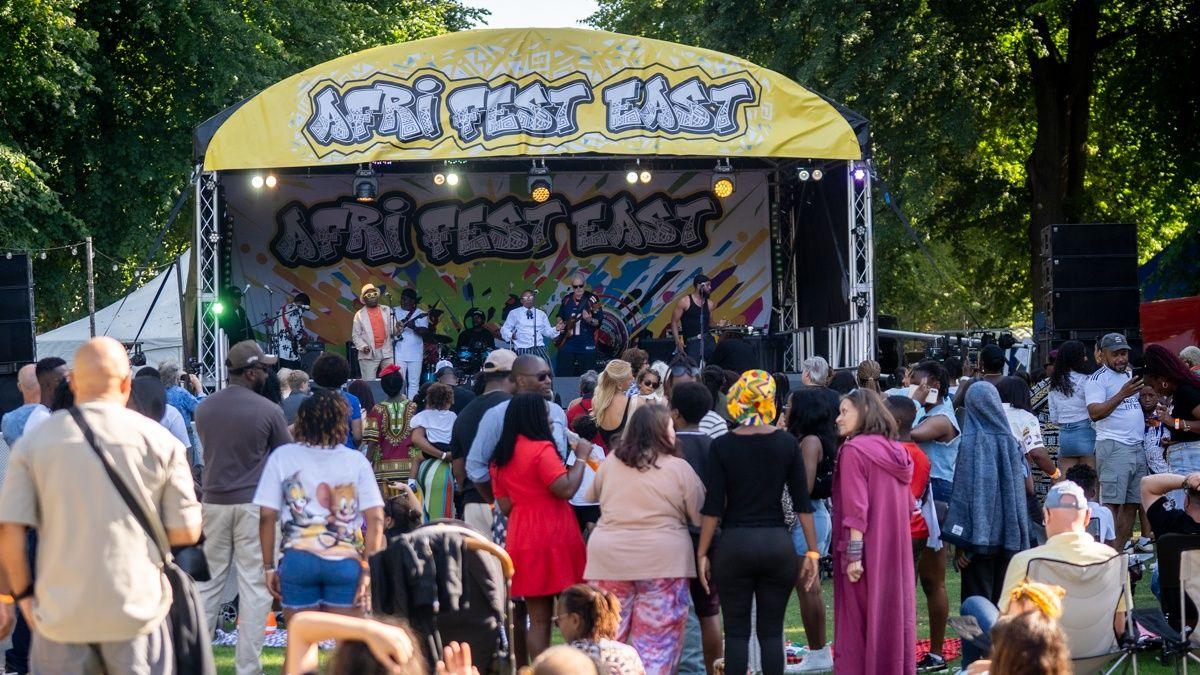 A festival stage in a park with a yellow banner with Afri-Fest East written in a graffiti style. Artists are performing on the stage and a crowd gathered in front.
