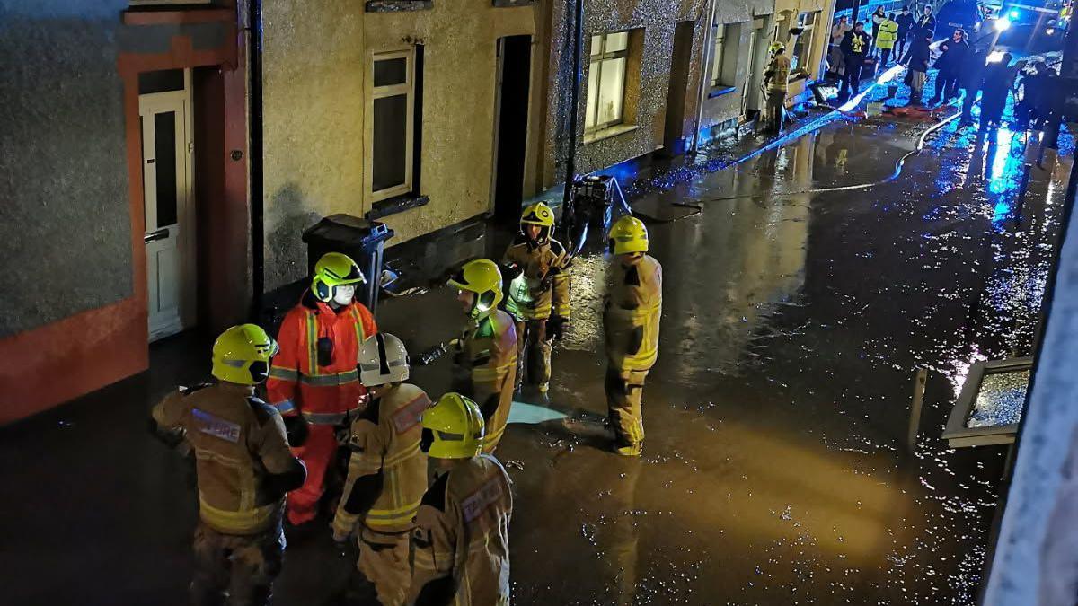 A fire crew stand in the middle of a street, their feet engulfed in sludge. Emergency vehicles are seen in the background as people look on.