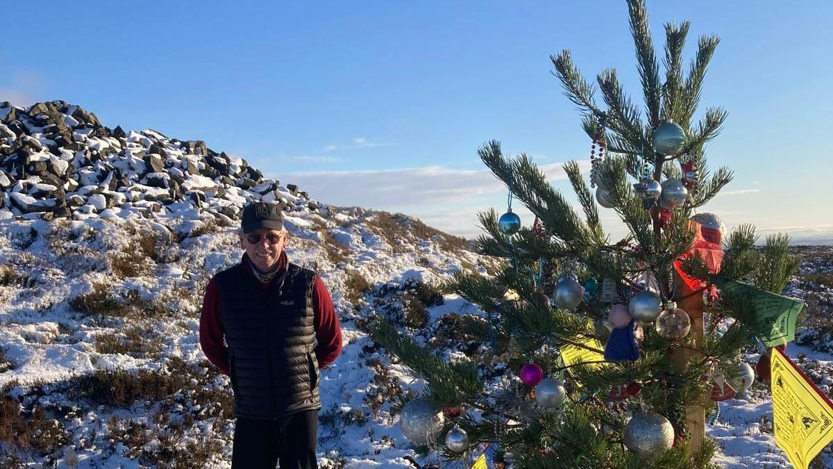 A man stands next to a Christmas tree on a hilltop with snow all around 