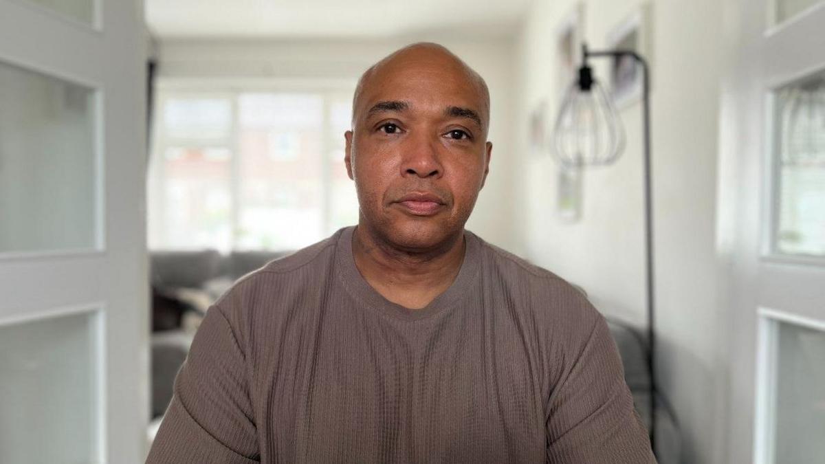 A profile shot of Kaine Fletcher's dad, Nathaniel Ameyaw, a Black man in his 40s, wearing a light brown T-shirt. He is facing the camera and two white doors are open behind him which lead into a living room. A black floor lamp can be seen on the right of the image behind Mr Ameyaw.