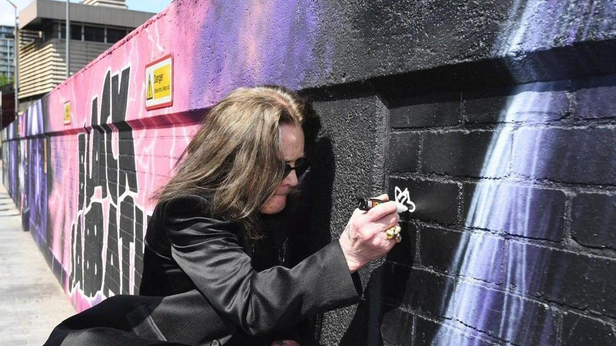 Ozzy Osbourne, an older man, in a black suit with long hair signing a street art mural. He is wearing black sunglasses and holding a white pen up to the wall. The mural is black, pink and purple and painted onto bricks in a city street.