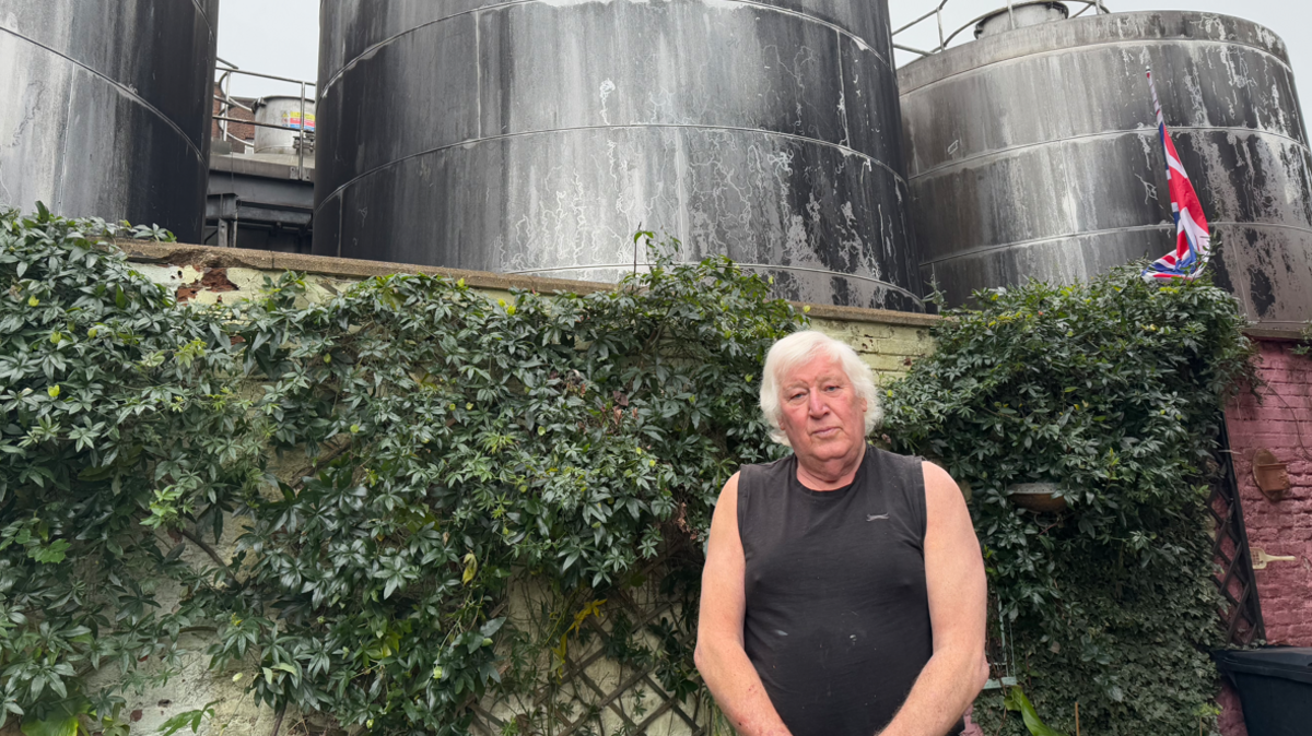 An elderly man wearing a black vest standing in front of a garden wall and a brewery