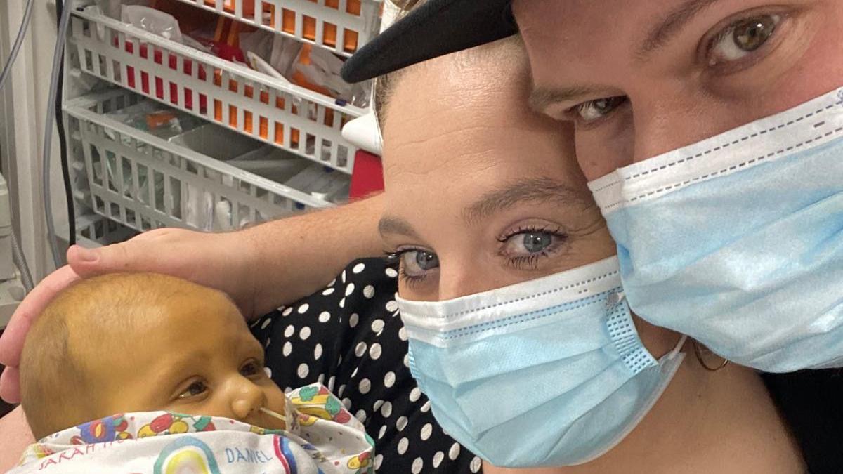 Close up selfie of Claire and Ollie, Leo's parents, wearing Covid masks and holding Leo, who is a small baby with yellow skin and a tube in his nose.