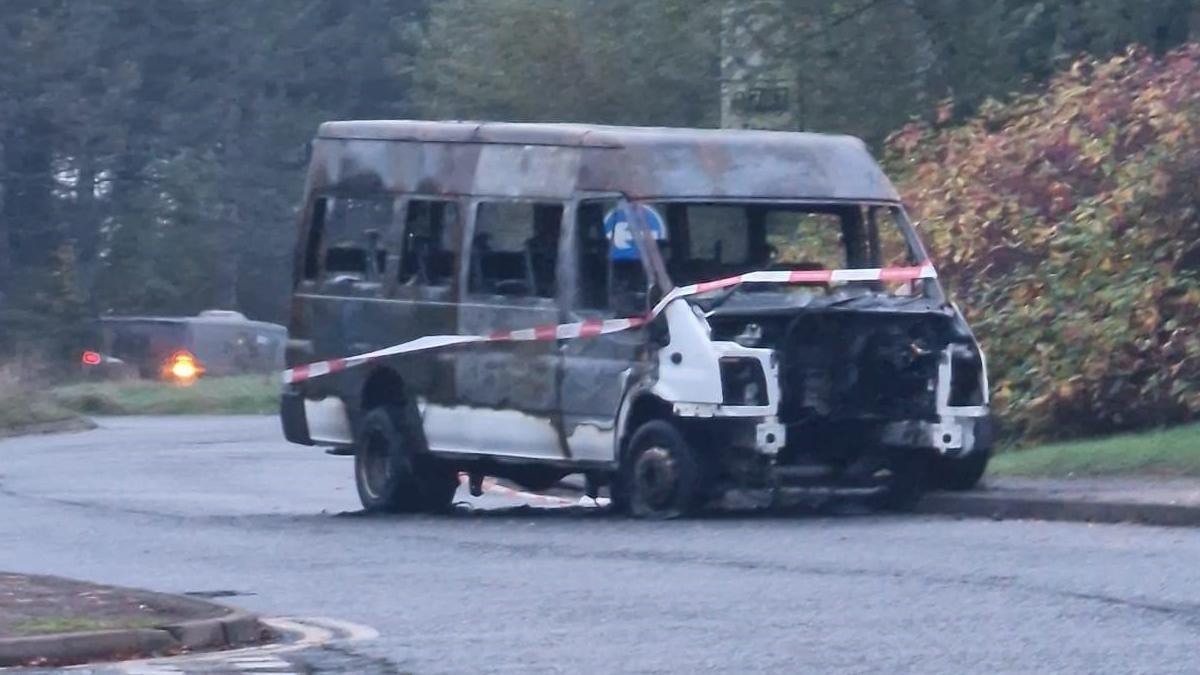 A burnt out bus, with no engine, with tape round it, on a road. A road sign is behind it and buses to the right. The bus was originally white in colour. 