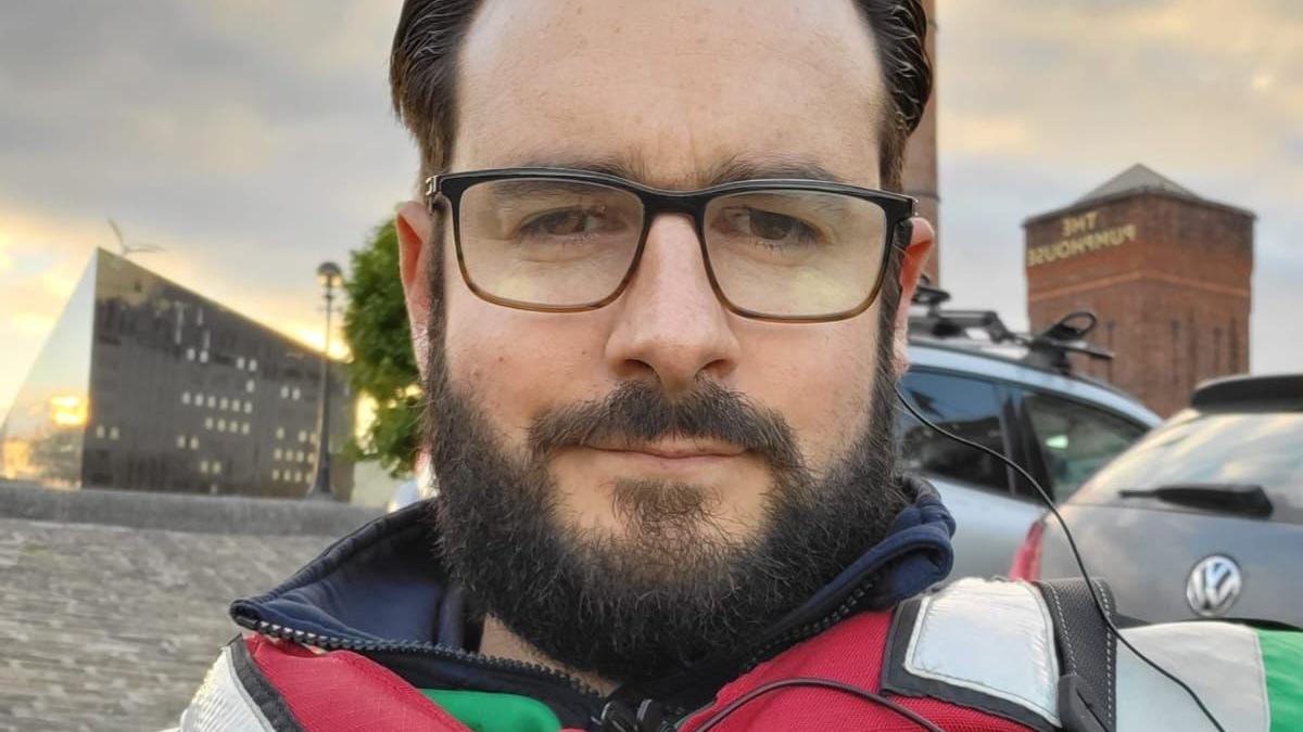 Lewis Bridson, who has black hair gelled back and a black beard, poses for a selfie in a red life-jacket with a view of Liverpool's waterfront in the background.