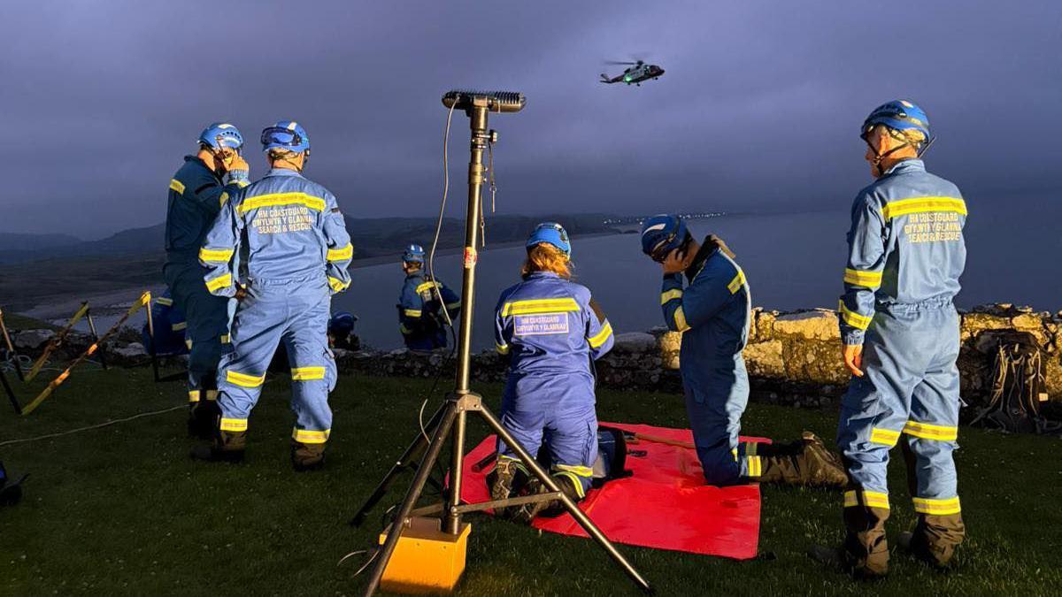 Nightime shot of RNLI crew on the cliffside. They were blue overalls with yellow stripes on them. A helicopter can be seen hovering above them. A tripod can be seen in the forefront of the image, illuminating the scene.