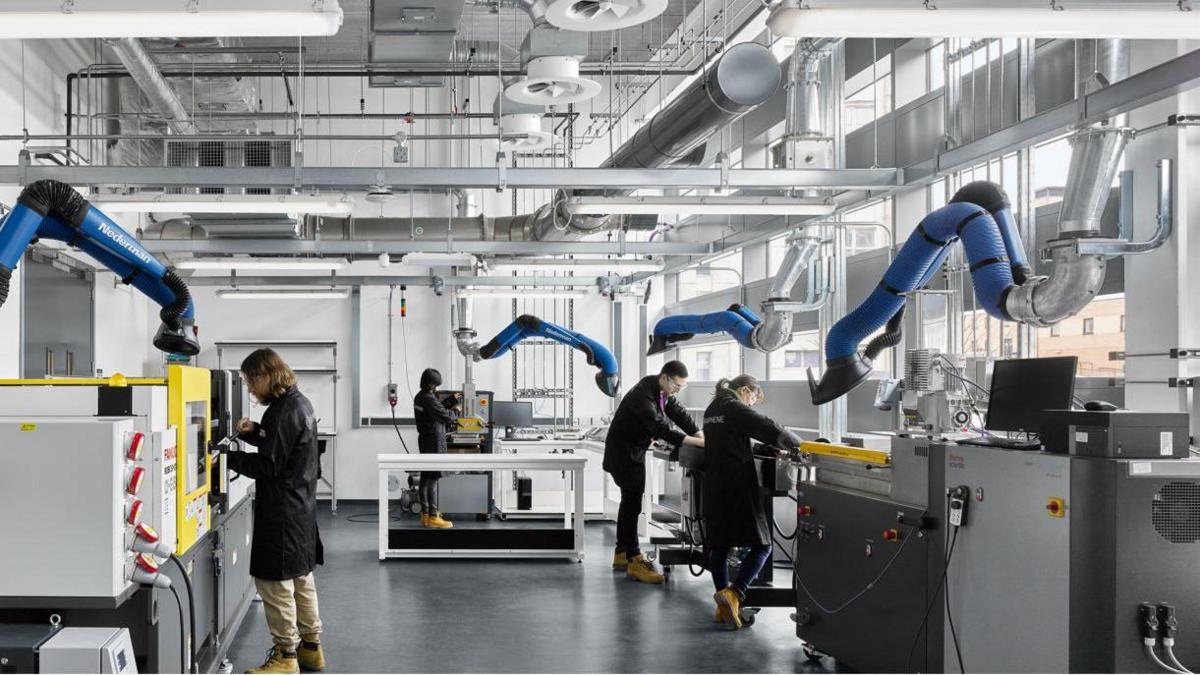 Lab workers wear black coats and goggles as they work in a lab at the Graphene Engineering Innovation Centre. Blue suction tubes can be seen overhead with various other pieces of scientific equipment seen. 