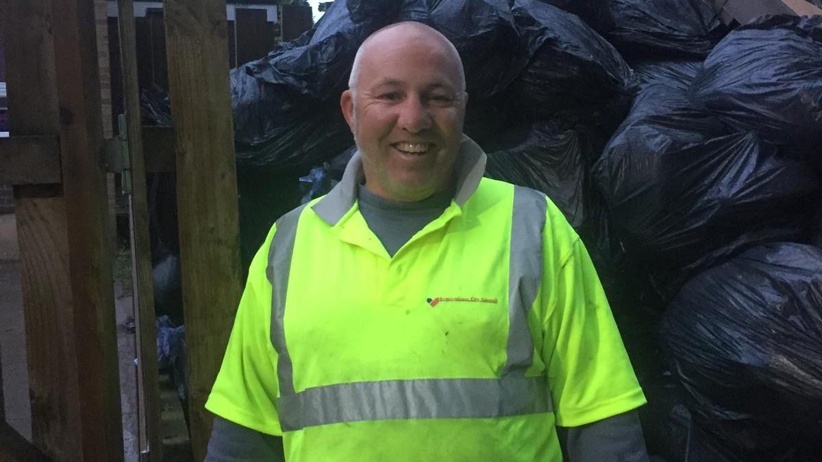 A man wearing a hi-vis jacket standing on front of a pile of black bin bags filled with rubbish.